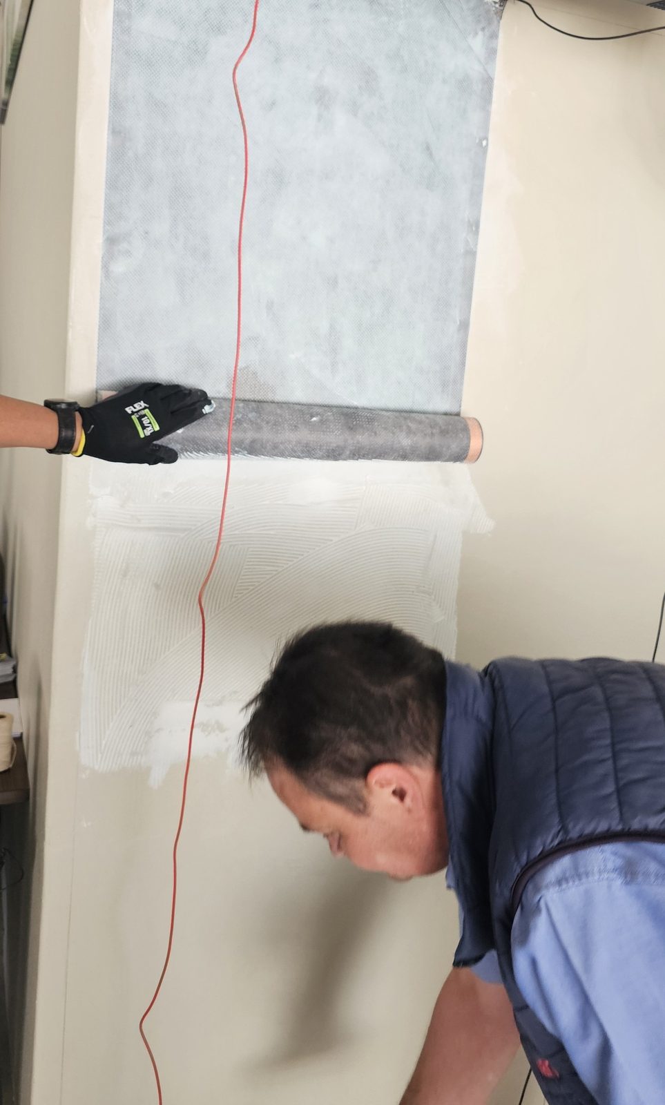 Synergy technician bonding a roll of carbon fibre heating film onto a wall with the adhesive combed on below it, the 48 V feed cable running down the wall